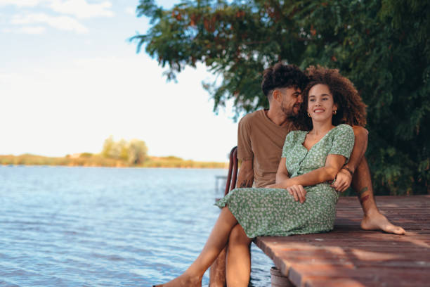 Romantic young couple sitting on a wooden pier, enjoying a sunny day while dipping their feet into the calm waters of a serene lake, embracing each other with smiles and laughter