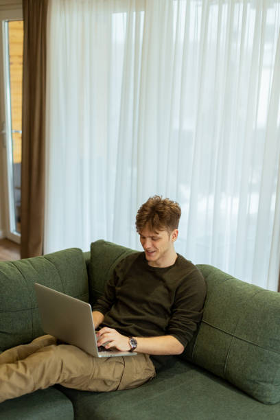 istock Man working diligently on his laptop while relaxing on a cozy green couch in a bright, airy living room 2196909422