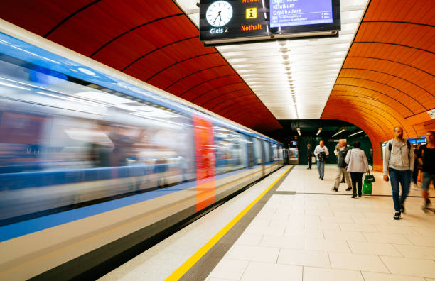 A fast-moving subway train leaving Marienplatz station with people on the platform under the station's signature orange walls and bright lighting. The motion blur of the train creates a dynamic and energetic urban scene.