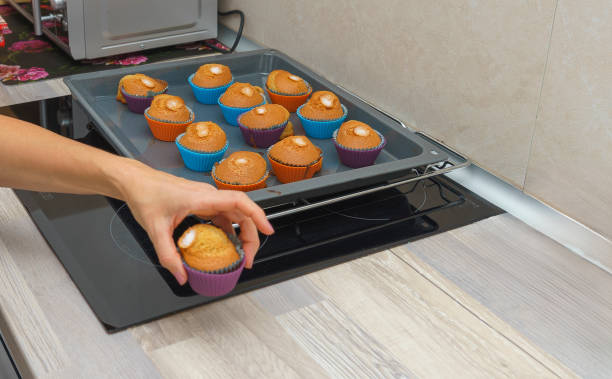 Close-up of person taking a freshly baked muffin from a baking tray. Golden brown muffins in colorful silicone molds cooling on the kitchen counter.