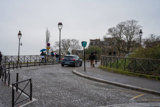 Paris,France-26.01.2025 Visitors stroll along a winding cobblestone path in Montmartre while a car navigates the narrow road. A cloudy sky adds a melancholic charm to the atmosphere.