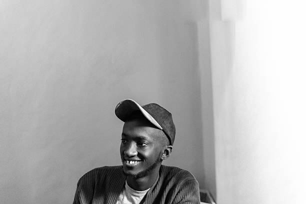 A black and white portrait of a young man wearing a cap, smiling warmly as he looks off-camera. The soft lighting and minimalist background highlight his expression, creating a timeless and introspective mood. EDITORIAL USE ONLY