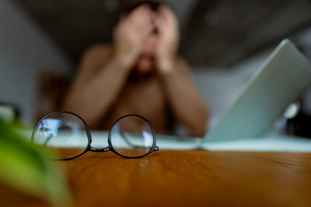 istock Eyeglasses resting on a desk with a stressed man in the background 2196936884