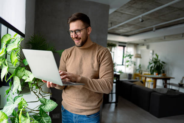 istock Young businessman working from home, standing and using laptop near window 2196937043