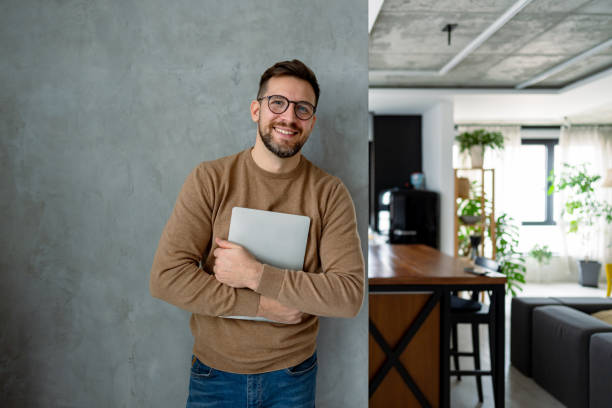 istock Young Man Smiling and Holding Laptop in Modern Apartment 2196937095