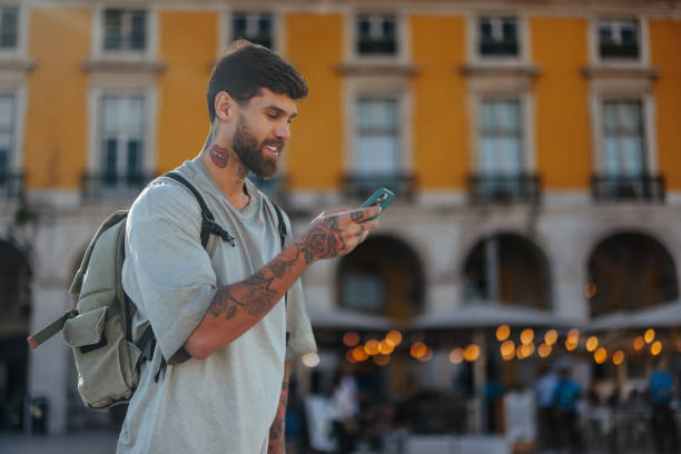 Young man with tattoos and backpack using mobile phone in commerce square, lisbon, portugal