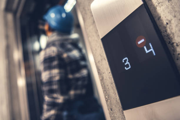 A construction worker in a hard hat stands by an elevator display indicating floors three and four during a busy workday.