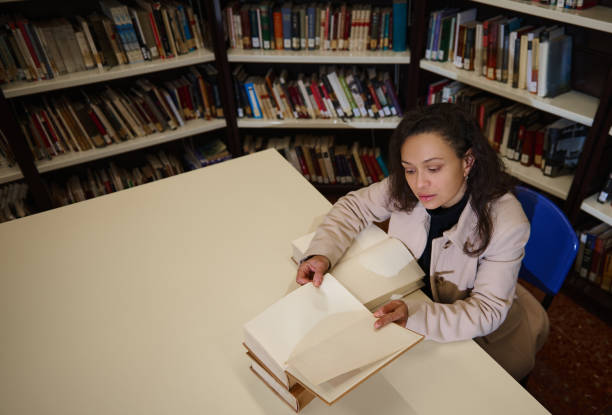 istock Woman reading at a library table surrounded by bookshelves for research 2196955147