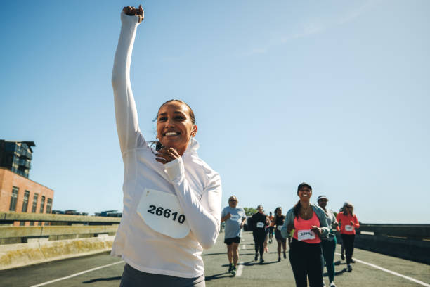 A joyful multiracial female runner celebrates her victory with her arm raised in triumph during a lively race event. Surrounded by other participants, she exudes a sense of achievement and motivation.