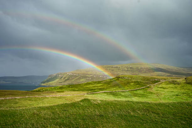 A breathtaking double rainbow arches over the rolling green hills of the Scottish Highlands, creating a stunning contrast against the dramatic stormy sky. Sunlight breaks through the clouds, illuminating the vibrant landscape and enhancing the natural beauty of this remote and peaceful setting.