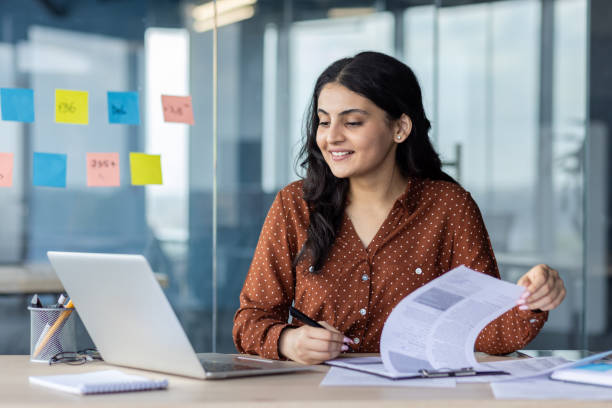 istock Young successful businesswoman working with documents inside the office. Office worker holding document in hands, filling out tax forms and reports using laptop 2196963475