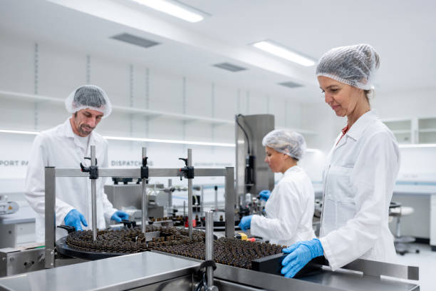 Assembly line worker in the production of pharmaceutical cosmetics. A female worker supplies the conveyor belt with bottles and closes the bottles with a drip cap, while a male worker supervises the operation of the conveyor belt and the line. Then the bottles are packed in the final packaging. The workers are dressed in white coats, protective gloves and caps.