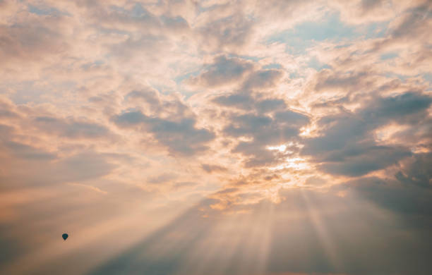 A breathtaking view of golden sun rays piercing through textured clouds, creating a dramatic and ethereal atmosphere over Cappadocia, Turkey. A lone hot air balloon drifts in the distance, adding a sense of adventure and tranquility to the scene. Cappadocia is known for its stunning landscapes, where ever-changing skies and soft light enhance the region's dreamlike charm