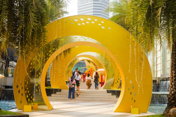Asian people and tourists between golden arches placed as chinese new year decor at shopping mall Siam Paragon. Steps are leading up from sidewalk alongside the mall. Steps are also decorated and flanked with palm trees. Some people are standing, others are walking up and others are walking down. At end of steps is small square and plateau between  mall and BTs station Siam. In background is next mall Siam Center. Mall is located at Phloen Chit Road in Bangkok downtown