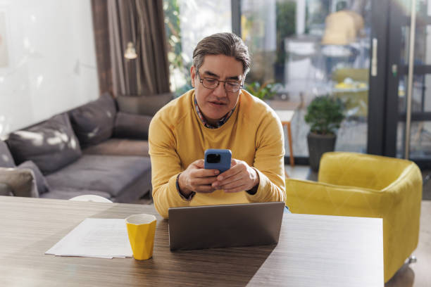 istock Mature man in a yellow sweater using smart phone and laptop at home 2196970134