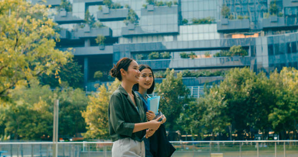 istock Two young Asian businesswomen discussing work while holds digital tablet and carries documents while walking outside office building. Sustainable business outdoor. 2196970422