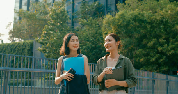 istock Two young Asian businesswomen discussing work while holds digital tablet and carries documents while walking outside office building. Sustainable business outdoor. 2196970438