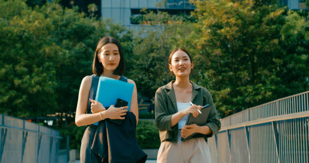 istock Two young Asian businesswomen discussing work while holds digital tablet and carries documents while walking outside office building. Sustainable business outdoor. 2196970444