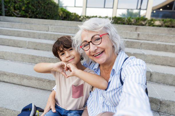 Happy grandmother and grandson are making heart shape with hands while taking a selfie sitting on urban steps