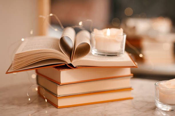 Burning scented candle saying on open paper book with folded pages in heart shape over glowing lights close up. Valentines Day.