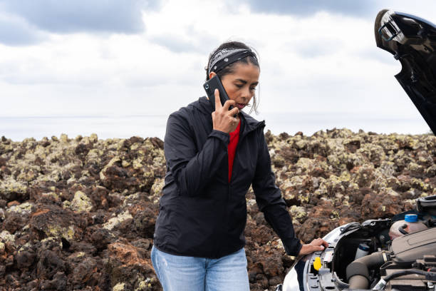 Frustrated female traveler standing near broken vehicle, volcanic coastline backdrop, awaiting roadside help with distressed expression