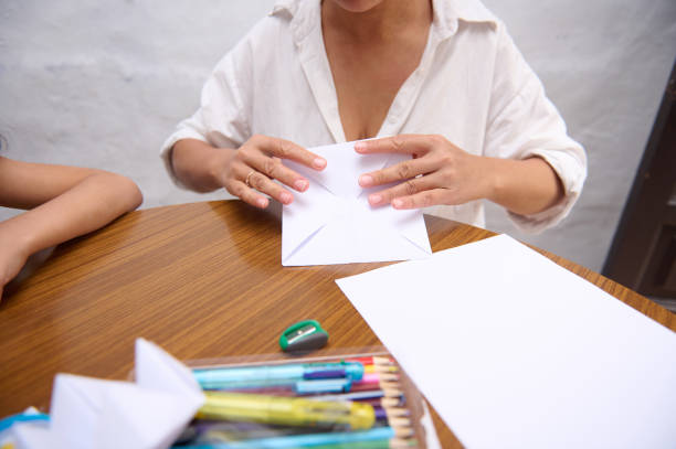 A person folding paper while engaged in a crafting activity on a wooden table. The setup includes school supplies like colored pens and pencils, suggesting a creative educational or hobbyist environment.