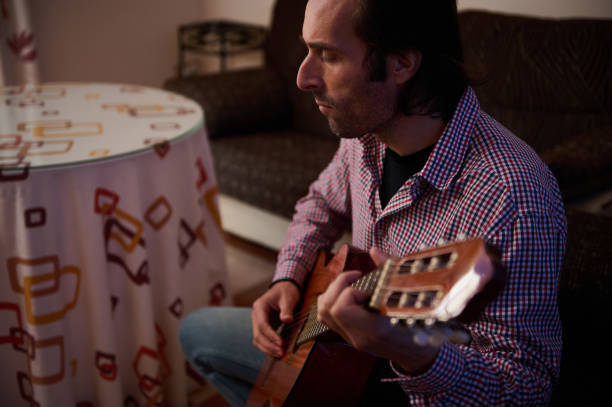 A close-up shot of a man seated indoors attentively playing an acoustic guitar, wearing casual clothing. The cozy ambiance adds to the image's calm and introspective mood.