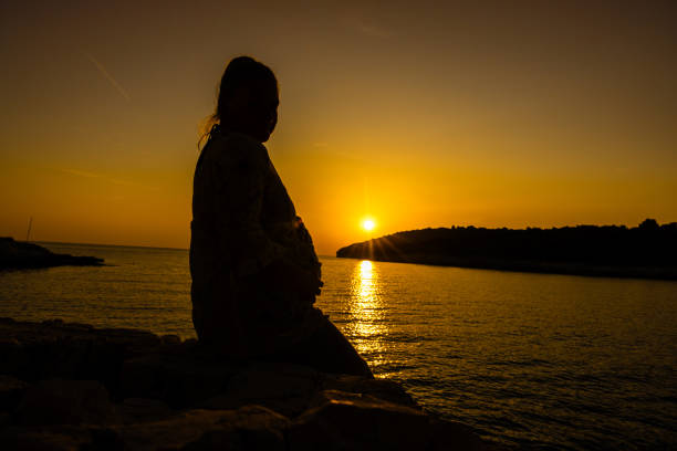 A serene photo of a mid-adult Caucasian pregnant woman by the sea, bathed in the glow of the setting sun. Her silhouette outlines her expecting figure against the tranquil backdrop.