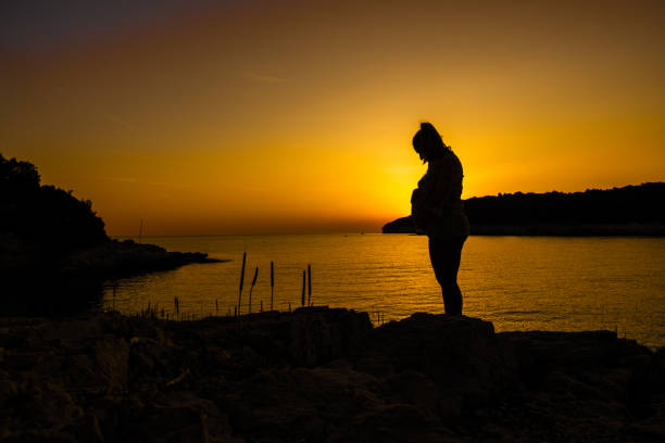 A pregnant mid adult Caucasian woman stands serene by the sea, silhouetted against an orange sunset sky, wearing casual clothes, reflecting on nature's beauty.