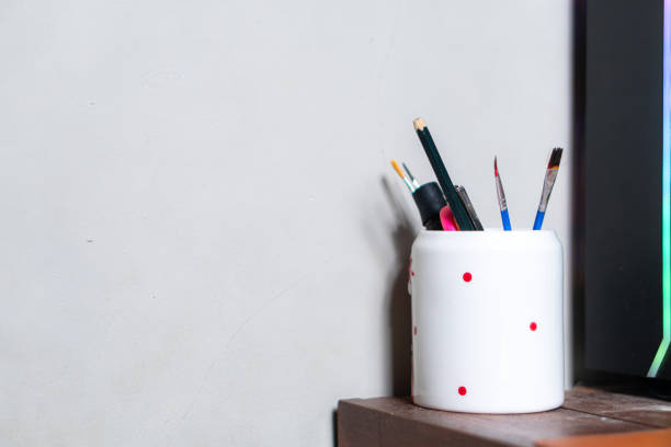 A white ceramic pot with red polka dots filled with various art supplies, including paintbrushes and pencils, placed on a wooden surface next to a wall.
