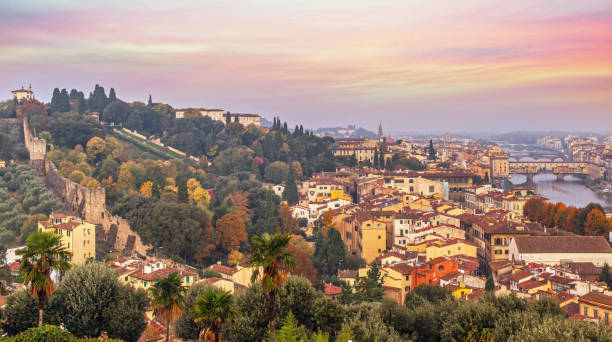 A breathtaking view of Florence at sunset, featuring historic city walls and vibrant rooftops leading to the iconic Ponte Vecchio. The lush greenery, autumnal trees, and warm hues of the sky create a picturesque scene, highlighting the city's timeless charm and architectural beauty.