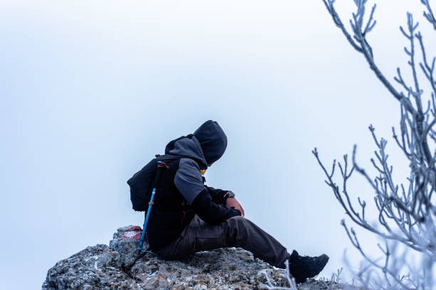 A hiker dressed for cold weather outdoors sits on a rocky surface surrounded by a snowy foggy environment. Depicting exploration, resilience, and the tranquility of nature. The muted colors and calm setting capture a peaceful moment of a wintertime journey in the wilderness.
