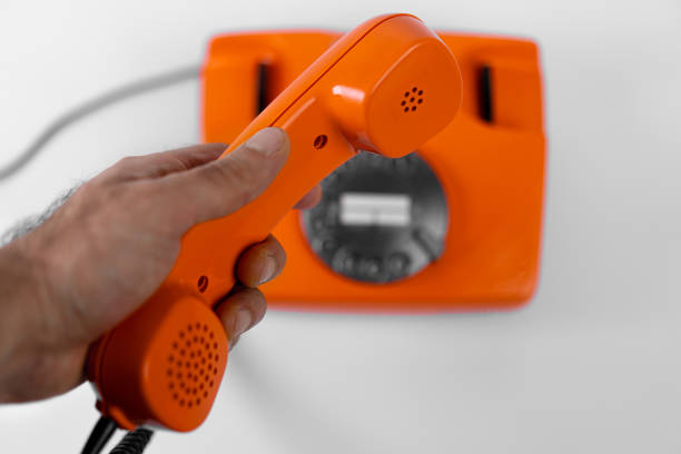 A hand grips a bright orange telephone receiver above a matching base while a person prepares to make a call. The setting is well-lit, enhancing the vibrant colors.