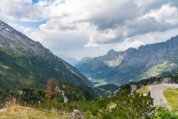 A mountain range in the Swiss Alps with a road running through it. The road is paved and has white lines. The mountains are covered in trees and the sky is cloudy