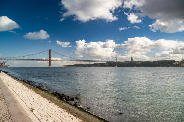 The iconic 25 de Abril Bridge stretches across the Tagus River under a vibrant sky, offering a serene moment along Lisbon’s scenic waterfront.