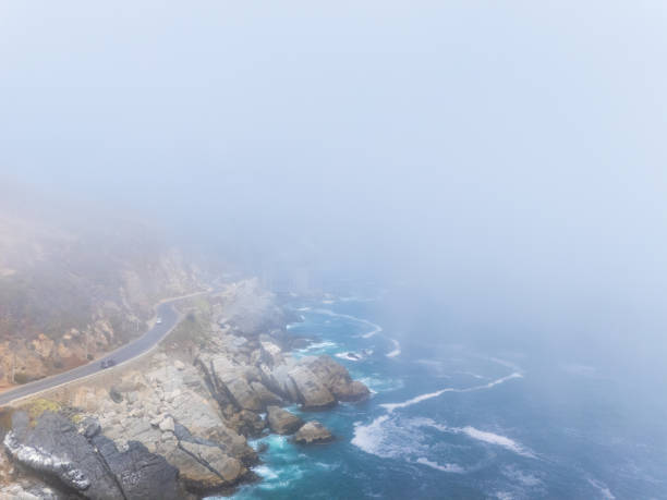 Misty coastal road winds along rocky cliffs by the ocean. Fog envelops scenic route, blending sea and sky.