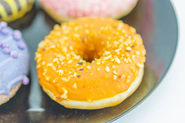Close-up of three donuts on a reflective surface, with the central one glossy orange and nutty, and others partially obscured The image has a shallow depth of field, bright lighting, and a plain whi