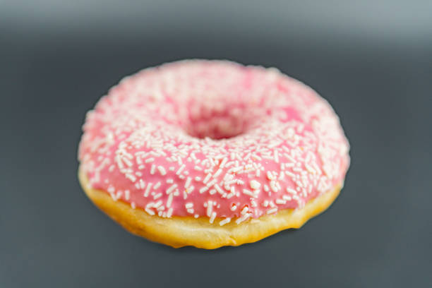 Stock photo showing elevated view of individual pink fondant iced ring doughnut on wood grain background.  The iced fried dough dessert is decorated with multicoloured hundred and thousand sugar sprinkles.