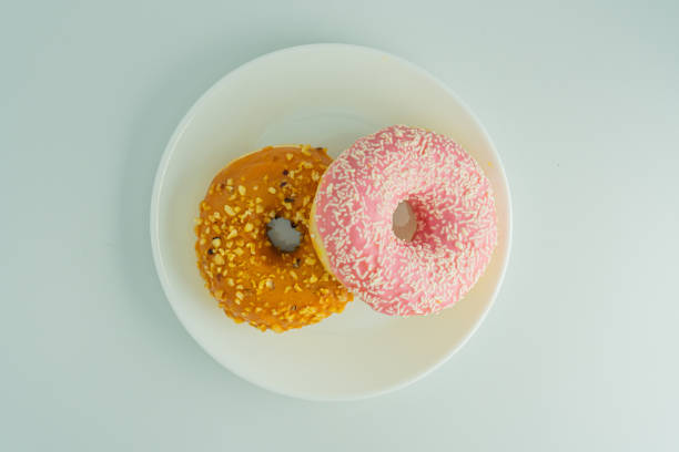 istock Two donuts on a white surface one yellow with scattered seeds or nuts, the other pink and sprinkled with white crunchy bits Ring-style, neutral background 2197088397