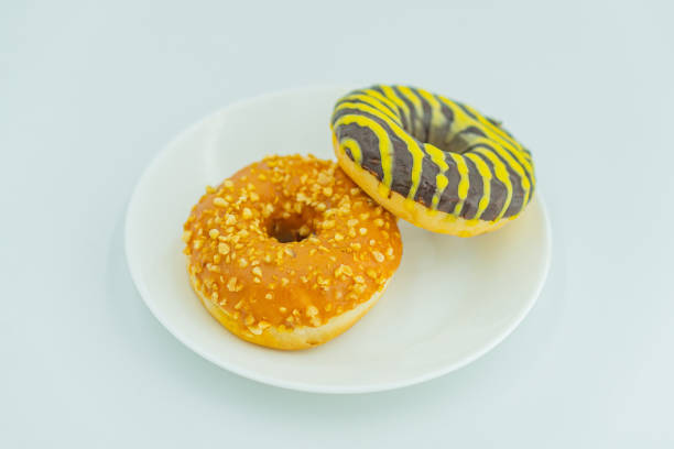 istock Image depicts two doughnuts on a white plate, one with glazeicing and nuts possibly almonds, the other with yellow icing and black stripes The backdrop is an indoor setting with a grayish color s 2197088442
