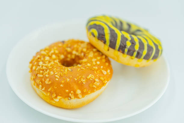 istock Two decorated donuts on a white plate against a light gray background The foreground donut is round, glazed, and covered with nuts possibly almonds or peanuts, while the one in the back has stripe 2197088448