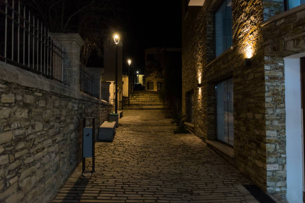 A narrow cobblestone alley at night, illuminated by warm and cool lights, with rustic stone walls and potted plants adding charm and mystery. Lefkara, Cyprus