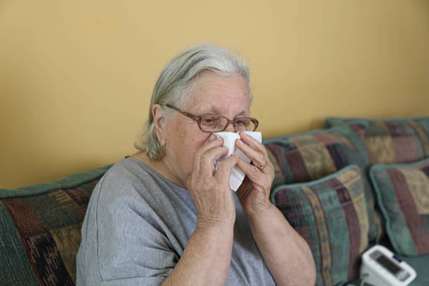 A close-up of an elderly woman with gray hair and glasses, holding a tissue to her nose. She appears unwell, sitting in a cozy home setting, conveying discomfort and illness.