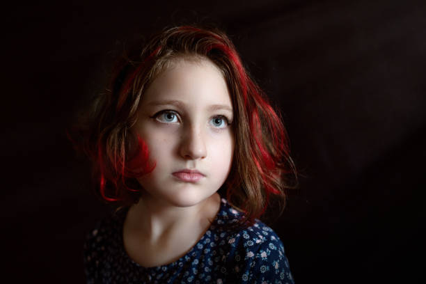 A contemplative young girl with striking blue eyes and vibrant red hair poses in a professional studio setting. Her upward gaze and thoughtful expression create an emotional atmosphere, while dramatic lighting techniques emphasize the contrast between light and shadow. Her patterned blue top adds a contemporary element to the fine art portrait.