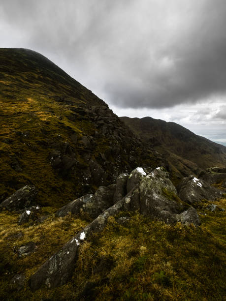 istock Grassy, rocky hillside leading up to a dark mountain ridge against a cloudy sky. 2197122128