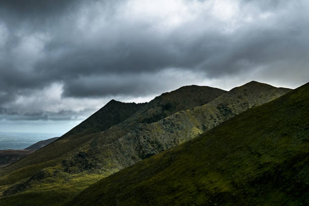 istock Sharp mountain peaks against a dark, stormy sky, lower slopes in shadow, hints of green vegetation. 2197122264
