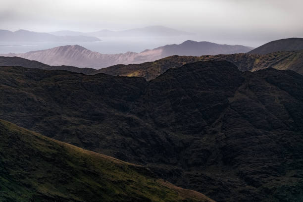 istock Layers of dark hills create a dramatic mountain landscape, shrouded in mist as the sun sets, revealing a distant coastline against a moody sky. 2197122267