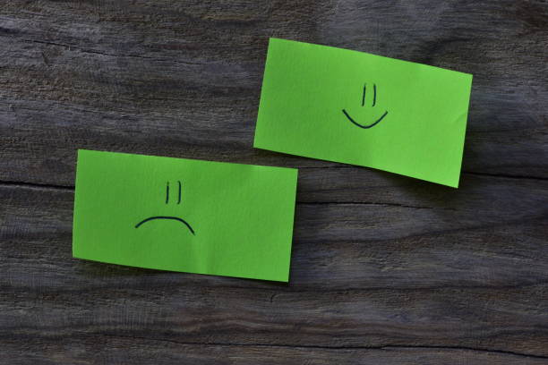 Smiley face and sad face on green sticky note on wooden board textured background. A pen draw smiley face and sad face on note paper.
