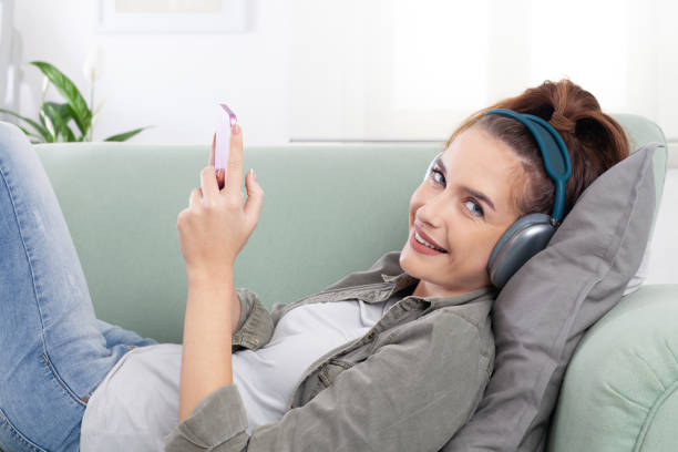 istock Young, relaxed, and smiling woman lying on a cozy couch, holding a smartphone while browsing messages, social media, music and digital content. She wears headphones, a modern, connected lifestyle 2197125527