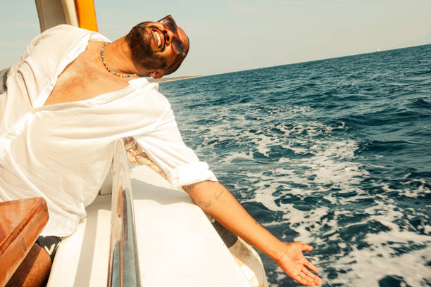 This image features a stylish, smiling man with a beard and sunglasses, wearing a white linen shirt and colourful beaded necklaces, sitting on a wooden boat during a sunny day at sea in Bodrum, Turkey. He is holding a smartphone and appears to be enjoying a relaxed moment, possibly taking a selfie or video calling. The background showcases the open ocean with gentle waves and a clear blue sky, with a distant coastline visible. The warm sunlight casts a golden glow on his face, enhancing the vacation, leisure, and travel vibes.